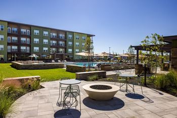A patio with a fire pit and chairs is surrounded by a grassy area and a building.